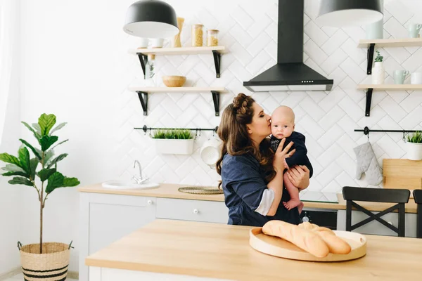 En vacker ung mamma sitter vid trä köksbordet och kysser hennes barn — Stockfoto