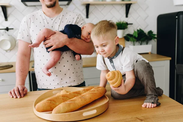 En liten pojke sitter på köksbordet och håller en croissant med sin far som håller en bebis bakom honom — Stockfoto