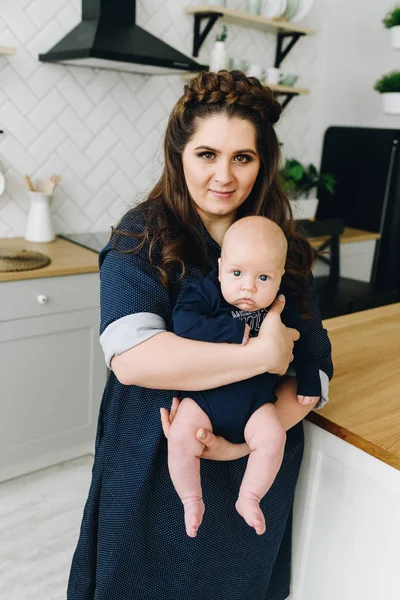 Una joven y hermosa madre de pie en la cocina y sosteniendo a su bebé — Foto de Stock