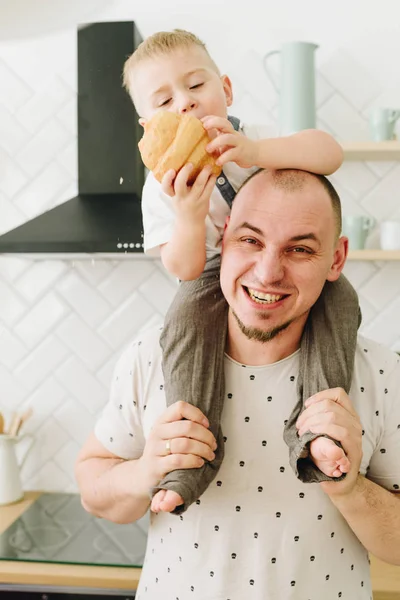 En söt liten kaukasisk pojke som äter en croissant medan hans far håller honom på axlarna — Stockfoto