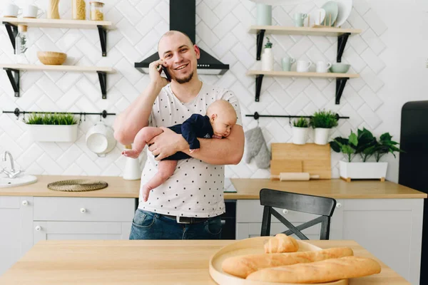 En ung far som håller sitt lilla barn och pratar på telefon i det lätta köket — Stockfoto