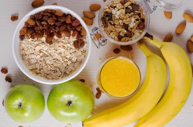 Sağlıklı kahvaltı. Beyaz kase kuru üzümlü yulaf ezmesi. İki yeşil elma, muz, fındık, granola. Düz üstten görünüm