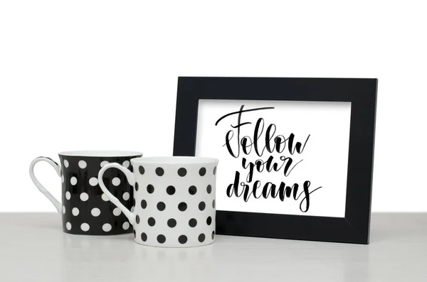 Follow your dreams. Handwritten text. Modern calligraphy. Black wooden frame for text. Black and white coffee Cup on the table. Close-up, white background