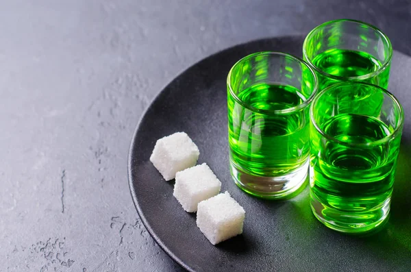 Absinthe green liquor in glasses. Alcoholic hallucinogenic beverage. Dark background. Pieces of white sugarin a plate . Copy space