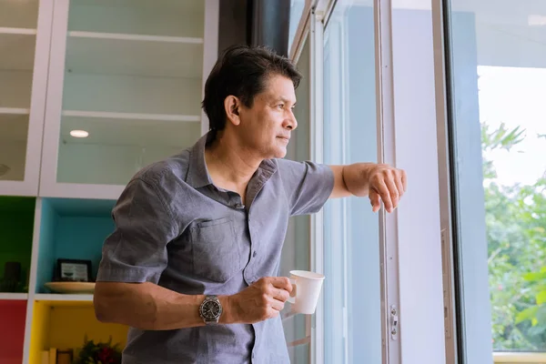 中年亚洲男子透过窗户看 喝着咖啡 用的想法 — 图库照片
