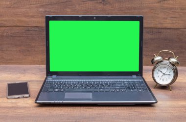 Front view of wooden office desktop with green screen notebook, alarm clock, mobile on wood wall background. Mock up