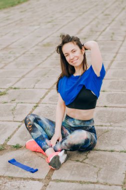 Koyu saçlı mavi saçlı kız gri asfalta oturur ve neşeyle gülümser, iyi günler.