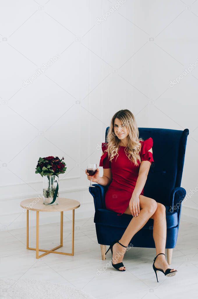 Portrait of a beautiful blonde girl in a red dress with a glass of red wine against the background of a stylish interior.