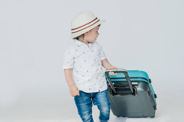 A boy with glasses and a hat is going to go on vacation, looks slyly from under his glasses. Leaning on a blue suitcase. The child is happy, smiling and laughing. Summer, vacation.