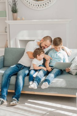 Mutlu genç baba dizüstü bilgisayar kullanarak kanepede oturuyor. Anaokulu çocuğu akıllı telefon tutarken rahatlıyor. Gülümsüyor baba ve küçük çocuk hafta sonunun keyfini çıkarıyor. Oğullarıyla mutlu bir baba.