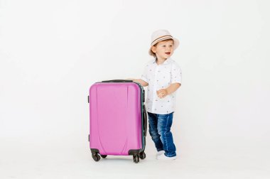 Gözlüklü ve şapkalı bir çocuk tatile çıkıyor, gözlüklerinin altından sinsi sinsi bakıyor. Mavi bir bavula yaslanarak. Çocuk mutlu, gülümsüyor ve gülüyor. Yaz, tatil.