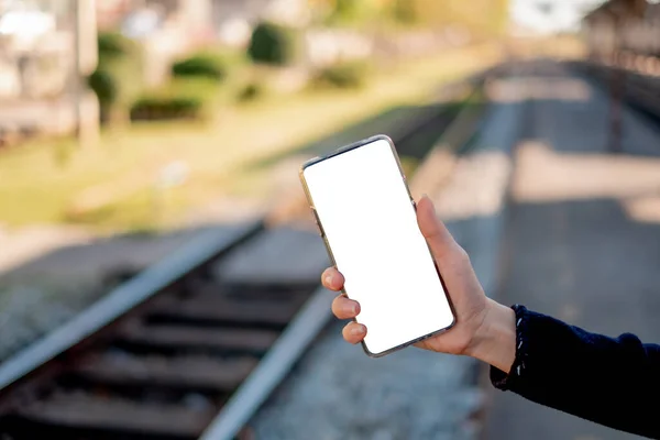 Woman\'s hand with a smartphone with a white screen. That has copy space for text space. With a Train tracks background.