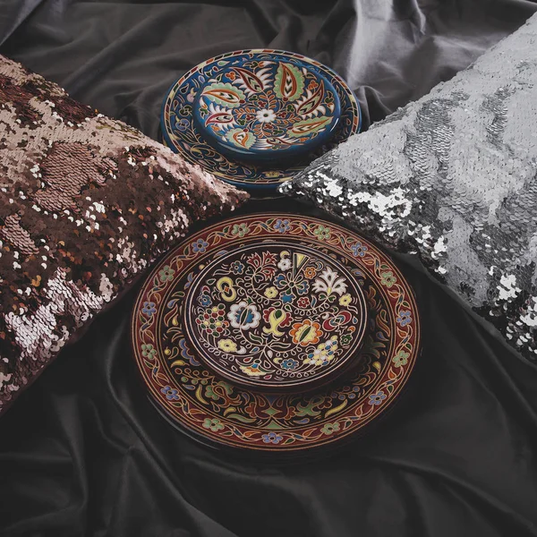Plates with ethnic ornament in composition with sequins pillows on a dark grey velvet fabric. Interior decoration concept.