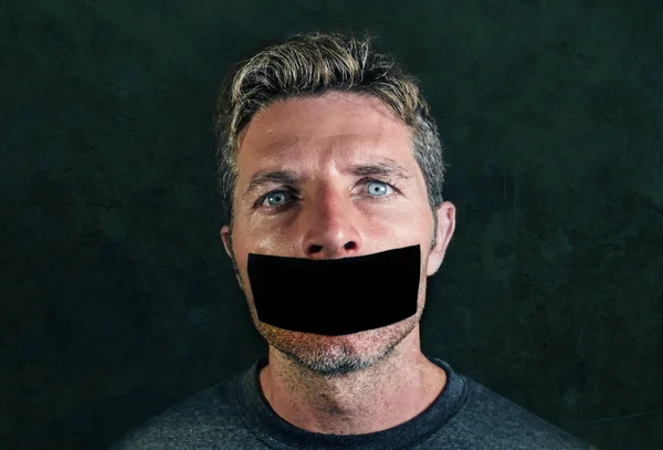 young man with mouth and lips sealed covered with adhesive tape in censorship coerced freedom of speech and forced silence and secrecy concept isolated on dark grunge background