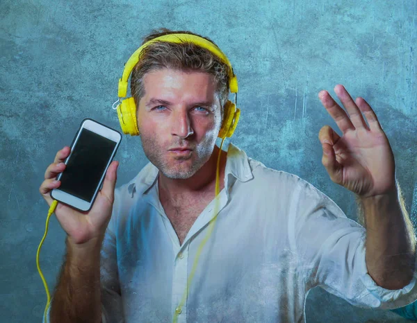 young attractive and happy cool man listening to music song with yellow headphones using internet mobile phone dancing in trance acting groovy and excited isolated on grey background