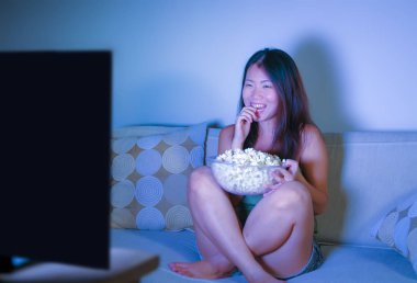 oturma odası kanepe oturma genç güzel ve mutlu Asyalı Koreli kadın kanepe izlerken televizyon komedi gösterisi geç gece yeme patlamış mısır gülümseyen ve yaşam tarzı kavramında gülüyor eğleniyor zevk