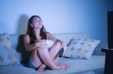 oturma odası kanepe oturma genç güzel ve mutlu Asyalı Koreli kadın kanepe izlerken televizyon komedi gösterisi geç gece yeme patlamış mısır gülümseyen ve yaşam tarzı kavramında gülüyor eğleniyor zevk