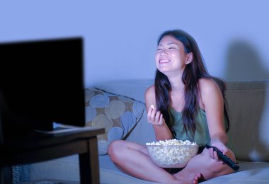 oturma odası kanepe oturma genç güzel ve mutlu Asyalı Koreli kadın kanepe izlerken televizyon komedi gösterisi geç gece yeme patlamış mısır gülümseyen ve yaşam tarzı kavramında gülüyor eğleniyor zevk