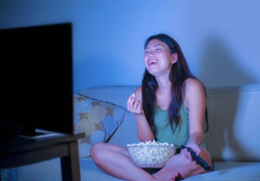 oturma odası kanepe oturma genç güzel ve mutlu Asyalı Koreli kadın kanepe izlerken televizyon komedi gösterisi geç gece yeme patlamış mısır gülümseyen ve yaşam tarzı kavramında gülüyor eğleniyor zevk
