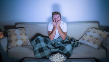 lifestyle portrait of attractive scared and nervous man watching suspense horror movie on television feeling stressed covering with blanket in panic sitting late night at home sofa couch clipart