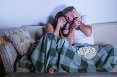 young mixed race attractive couple with Asian Korean woman and white man enjoying together watching television horror movie scared girlfriend in panic covering eyes on sofa couch eating popcorn  clipart