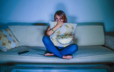 young beautiful and scared woman watching horror or suspense movie eating popcorn sitting at home sofa couch late night in panic and fear face expression in girl domestic lifestyle concept clipart