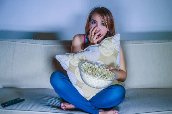 geç gece kanepede yastık tutan panik ve sürpriz yüz ifadesi içinde genç güzel ve korkmuş latin kadın kanepe evde oturan patlamış mısır yemek korku ya da gerilim film izlerken