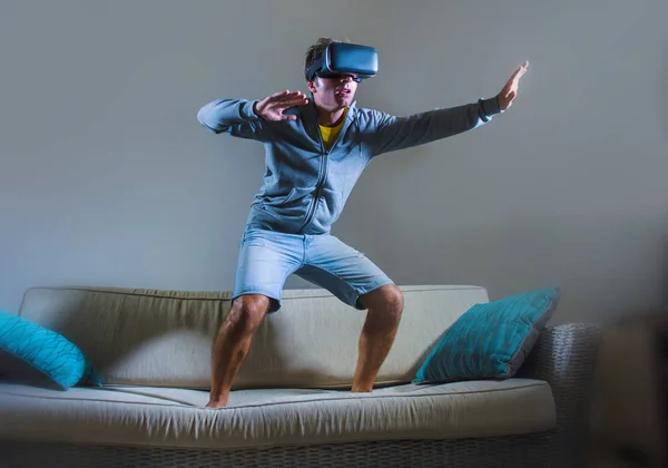 young attractive gamer man using VR goggles headgear technology playing simulator 3D video game having fun on home sofa couch enjoying exciting experience playing with virtual reality glasses