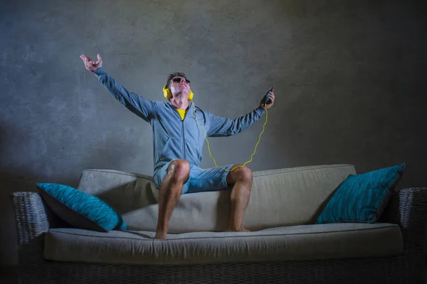 young attractive and cool happy man listening to techno music with yellow headphones jumped on top of home sofa couch dancing internet song excited in trance holding mobile phone