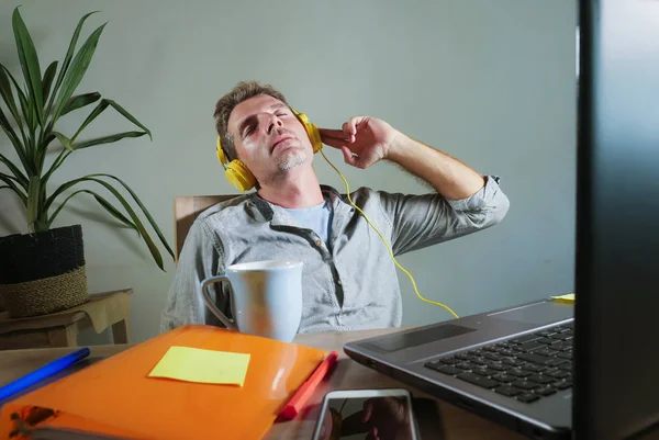 young attractive and happy man with yellow headphones sitting at home office desk working with laptop computer having fun listening to music enjoying song excited in lifestyle concept