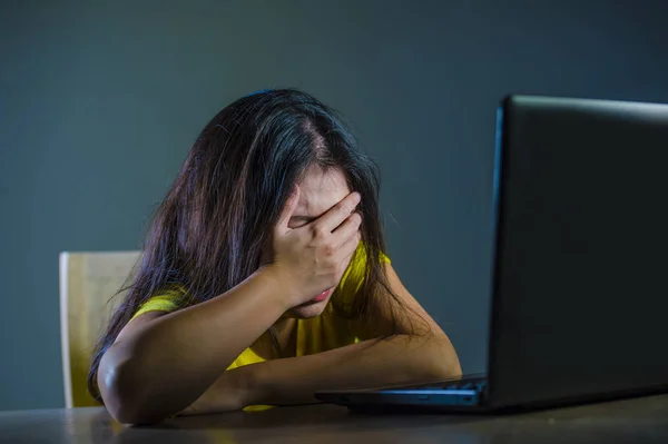 dramatic portrait of young sad and scared woman covering face with hands stressed and worried looking at laptop computer isolated on dark background in cyber bullying and internet problem