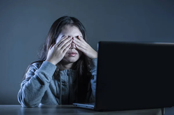 dramatic portrait of young sad and scared woman covering face with hands stressed and worried looking at laptop computer isolated on dark background in cyber bullying and internet problem