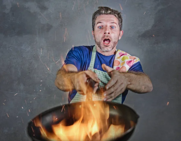 young attractive and shocked messy home cook man with apron holding pan in fire burning the food in kitchen disaster and unskilled and unexperienced terrible home cook at domestic cooking