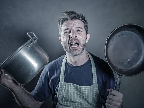 young stressed and funny lazy man with apron holding kitchen pan and kitchen pot screaming in stress desperate crying isolated background in domestic cooking work and messy husband