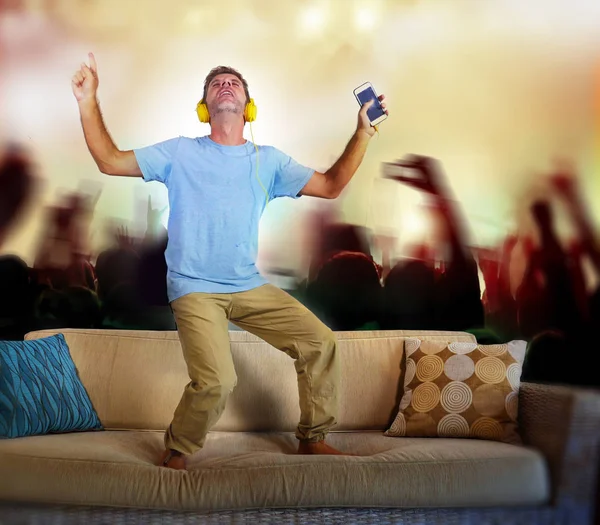 young happy and excited man jumping on sofa couch listening to music with mobile phone and headphones singing and dancing crazy imagine as famous rock band concert and fans audience