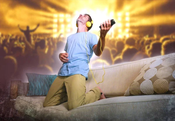 young happy and excited man jumping on sofa couch listening to music with mobile phone and headphones playing air guitar crazy having fun at home living room in audio fan lifestyle concept