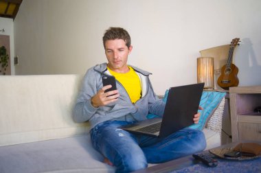 dizüstü bilgisayar ve cep telefonu freelancer iş ve bağımsız iş yaşam tarzı mutlu ağ kanepede oturan ev oturma odasından çalışan genç çekici ve başarılı erkeğin 