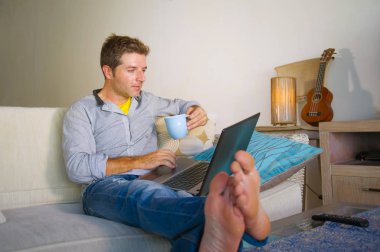 çevirmen ve bağımsız iş iş kavramı mutlu kahve içme dizüstü bilgisayarda ağ kanepede oturan ev oturma odasından çalışan genç çekici ve başarılı erkeğin