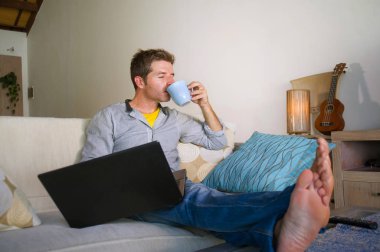 çevirmen ve bağımsız iş iş kavramı mutlu kahve içme dizüstü bilgisayarda ağ kanepede oturan ev oturma odasından çalışan genç çekici ve başarılı erkeğin