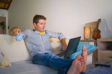 çevirmen ve bağımsız iş iş kavramı mutlu kahve içme dizüstü bilgisayarda ağ kanepede oturan ev oturma odasından çalışan genç çekici ve başarılı erkeğin