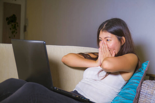 lifestyle portrait of young beautiful Asian Chinese student woman happy and relaxed at home sofa couch using internet on laptop computer networking looking surprised and amazed