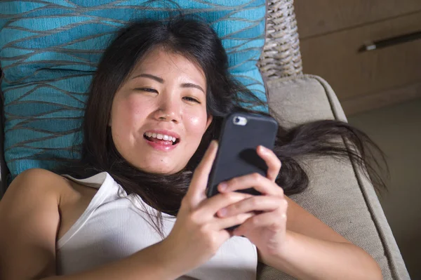 young sweet happy and pretty Asian Chinese woman using internet social media app on mobile phone smiling cheerful lying on home sofa couch in online communication lifestyle concept