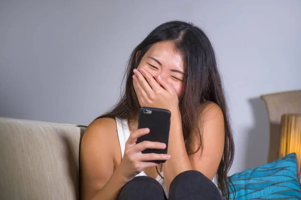 young sweet happy and pretty Asian Korean girl using internet social media app on mobile phone laughing cheerful having fun at home sofa couch in online communication lifestyle concept