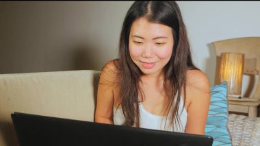 Genç güzel Asyalı Koreli öğrenci kız mutlu ve evde rahat koltuk kanepe üzerinde dizüstü bilgisayar ağ online teknoloji iletişim yaşam tarzı kavramı içinde neşeli internet kullanımı 