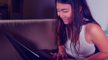 Genç güzel Asyalı Koreli öğrenci kız mutlu ve evde rahat koltuk kanepe üzerinde dizüstü bilgisayar ağ online teknoloji iletişim yaşam tarzı kavramı içinde neşeli internet kullanımı 