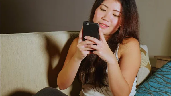 young sweet happy and pretty Asian Korean woman using internet social media app on mobile phone smiling cheerful having fun at home sofa couch in online communication lifestyle concept