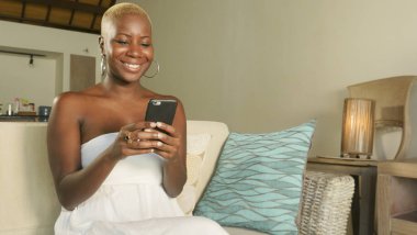 Genç güzel ve mutlu internet sosyal medya app üstünde hareket eden ağ ve online evde oturma odası koltuk kanepe oturup kalma telefon kullanarak siyah Afrikalı-Amerikalı kadın gülümseyen heyecan