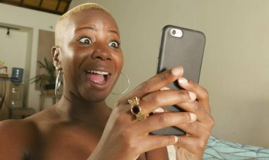 Genç güzel ve mutlu internet sosyal medya app üstünde hareket eden ağ ve online evde oturma odası koltuk kanepe oturup kalma telefon kullanarak siyah Afrikalı-Amerikalı kadın gülümseyen heyecan