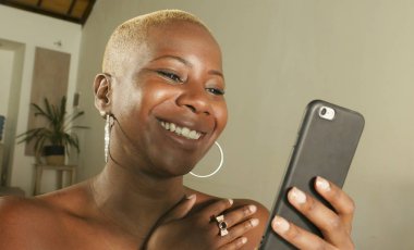 Genç güzel ve mutlu internet sosyal medya app üstünde hareket eden ağ ve online evde oturma odası koltuk kanepe oturup kalma telefon kullanarak siyah Afrikalı-Amerikalı kadın gülümseyen heyecan