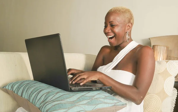 自宅のラップトップ コンピューターがブロガーやインフルエンサーのライフ スタイルのリラックスしたリビング ルームのソファにソーシャル メディアを使用してインターネット上で若い美しい 幸せな黒アフロ アメリカ人女性興奮して笑みを浮かべて楽しんでください — ストック写真